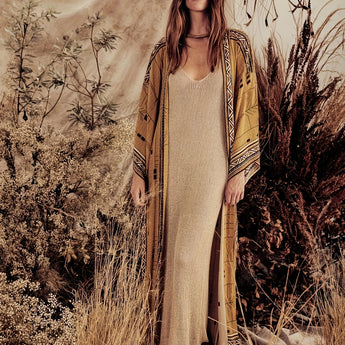 A woman with long hair wearing a beige bohemian kimono with a tribal pattern, standing in a grassy area surrounded by plants, embodying boho-chic desert fashion.