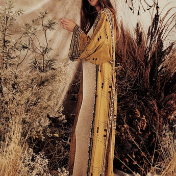 A woman wearing a Bohemian beige kimono with a boho-chic tribal pattern, standing in a sunlit grassy field, showcasing the outerwear's long sleeves and waist design.