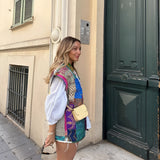 Bohemian embroidered patchwork dress being worn by a model standing near a window. The dress features intricate embroidery, a relaxed fit, and vibrant patchwork colors, embodying artisan summer fashion. The model's attire includes stylish eyewear and showcases the dress's detailed shoulder and sleeve design, with a focus on the waist and knee-length hemline.