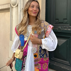 A woman wearing a bohemian embroidered patchwork dress, featuring a mix of colorful patterns, standing in a stylish pose. She accessorizes with a necklace, eyewear, and holds a small bag, embodying a chic summer fashion look. Her smile and confident stance highlight the dress's design, including its distinctive shoulder and sleeve details.