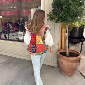 Bohemian embroidered patchwork dress displayed on a model standing in a relaxed pose, showcasing intricate textile patterns. The dress features short sleeves and a fitted waist, with houseplants and flowerpots in the background enhancing the artistic, summery vibe.