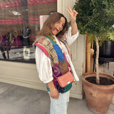Bohemian embroidered patchwork dress with colorful patterns, featuring flowing sleeves and a defined waist, styled for summer fashion. The model is standing outdoors, wearing sunglasses, near a houseplant and flowerpot.