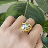 A hand adorned with a bold gold adjustable statement ring featuring a large reflective gemstone on the finger.