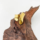 A bold gold statement ring with a modern, sculptural design, displayed against a natural wood background with tree trunks and twigs.