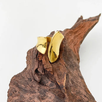 A bold gold statement ring with a modern, sculptural design, displayed against a natural wood background with tree trunks and twigs.