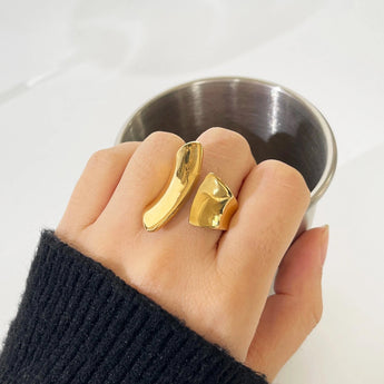 A hand wearing a bold gold statement ring with a modern, sculptural design on the finger, surrounded by greenery.