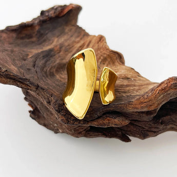 Modern sculptural bold gold statement ring displayed on natural wood with plants in the background.