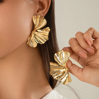 Oversized gold leaf statement earrings with bold textured botanical design, worn on ears and partially displayed by a hand showcasing the intricate details and shiny gold color.