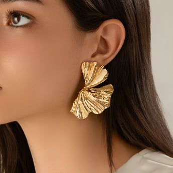 Oversized botanical earrings in gold leaf design, worn on ear with textured details, showcasing bold statement accessory against skin and shoulder background.