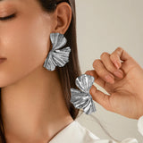A close-up of a woman wearing oversized botanical statement earrings with a bold textured gold leaf design. The earrings are displayed against her skin, near her ear and neck, with her black hair partially visible.