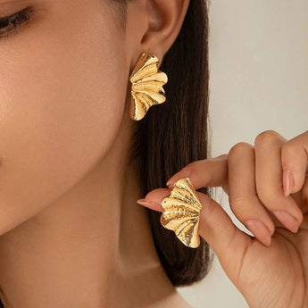 A close-up of a woman's ear adorned with bold, textured gold leaf statement earrings, highlighting designer fashion jewelry in gold color, with visible skin, eyelashes, and fingers gently touching the earring.