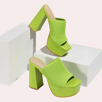 A pair of bright orange ankle boots with stretch fit sock heels featuring neon details, worn by a person standing on a floor.