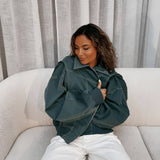 
A woman wearing a green oversized leather bomber jacket, styled for trendy fall fashion, sitting comfortably on a couch. The jacket features long sleeves, a relaxed fit around the waist, and pairs with her casual look.
