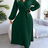
A woman wearing a green pleated midi wrap dress with an elegant V-neck and tie waist, styled with long sleeves, standing outdoors surrounded by plants.