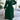 
A woman wearing a green pleated midi wrap dress with an elegant V-neck and tie waist, styled with long sleeves, standing outdoors surrounded by plants.