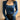 
A woman wearing a blue ribbed knit top featuring a square neckline and long sleeves. The sweater fits snugly, highlighting the shoulders and waist.