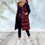 Woman wearing a burgundy long puffer coat with a detachable hood, standing and smiling. The coat features a fur-lined hood, long sleeves, and extends down to the knees. The model has a happy expression, showcasing the coat's stylish and warm design.
