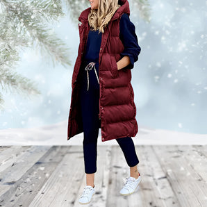 Woman wearing a burgundy long puffer coat with a detachable hood, standing and smiling. The coat features a fur-lined hood, long sleeves, and extends down to the knees. The model has a happy expression, showcasing the coat's stylish and warm design.