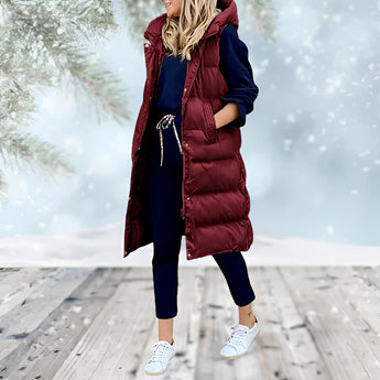 Woman wearing a burgundy long puffer coat with a detachable hood, standing and smiling. The coat features a fur-lined hood, long sleeves, and extends down to the knees. The model has a happy expression, showcasing the coat's stylish and warm design.