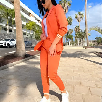 A woman with black hair wearing a taupe blazer and brown trousers, styled with sneakers, standing under a clear sky. She is accessorized with sunglasses, embodying a business casual look. Her outfit is in the orange color variant, size 2XL.