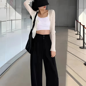 A woman dressed in black wide-leg trousers, a cropped tank top, and a cardigan, accessorized with a sun hat and sunglasses, showcasing a chic urban fashion style.