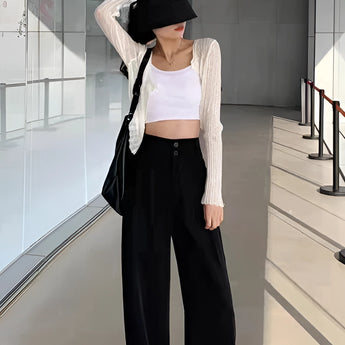 A woman dressed in black wide-leg trousers, a cropped tank top, and a cardigan, accessorized with a sun hat and sunglasses, showcasing a chic urban fashion style.