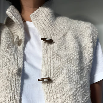 Chunky cable knit vest with wool toggle buttons, featuring a grey color and textured design, styled over a dress shirt.