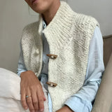 A grey chunky cable knit vest with wool toggle buttons, featuring textured patterns, shown over a white dress shirt with visible sleeves and shoulder details.