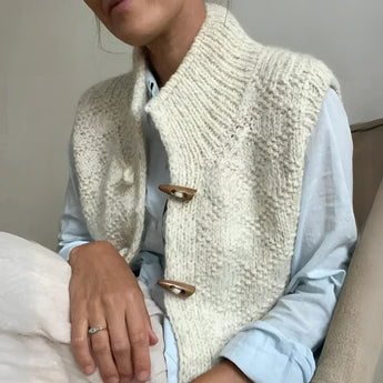 A grey chunky cable knit vest with wool toggle buttons, featuring textured patterns, shown over a white dress shirt with visible sleeves and shoulder details.