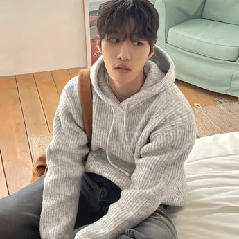 Oversized gray hoodie with a chunky knit design, suitable for winter wear, featuring long sleeves and a relaxed fit, designed for unisex use, with a person sitting while wearing it.