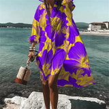 Colorful retro print kaftan in vibrant psychedelic patterns, primarily in purple and violet hues, designed as a beach cover-up. The kaftan is modeled by a person standing against a backdrop of azure sky and beach scenery, emphasizing a relaxed and stylish look ideal for travel and beachwear.