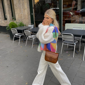 A woman wearing a colorful oversized patchwork cardigan and sunglasses is sitting on a chair near a window, with plants visible in the background.