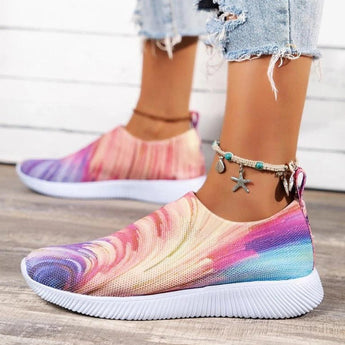 Colorful women's slip-on walking sneakers with a rainbow mesh design, shown on a model wearing jeans. The lightweight sneakers, size 42, feature vibrant hues and are designed for comfort and style.