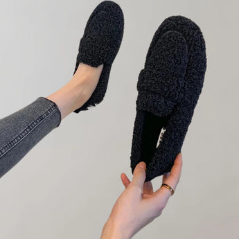 A pair of light grey cozy fleece slippers for women, featuring a soft plush material and anti-slip soles, displayed in a casual indoor setting.