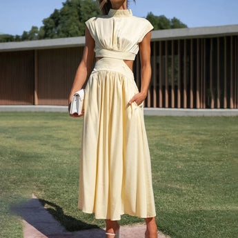 Elegant black midi dress with back cutout, worn by a model outdoors. The dress features a sleek one-piece design with sleeves, highlighting the neckline and waist. The model's hair flows naturally, complementing the elegant evening wear look against a subtle sky backdrop with greenery.