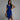 
A woman smiling while wearing a blue sleeveless V-neck wrap cocktail dress that is knee-length. The dress is form-fitting, highlighting its sheath style, and the woman has her hand placed near her waist, showcasing the elegant design. Her hairstyle complements the sophisticated look of the dress.