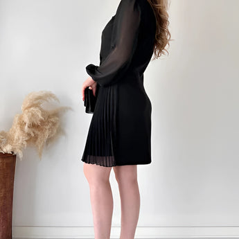 Elegant black pleated mini dress featuring long sleeves, worn by a woman standing upright. The dress is styled with an office chic look, highlighting the shoulder, waist, and knee areas.