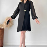 Elegant black pleated long sleeve mini dress styled with chic office wear, featuring a model wearing black footwear. The dress is a one-piece garment with a knee-length hem, suitable for a professional setting.