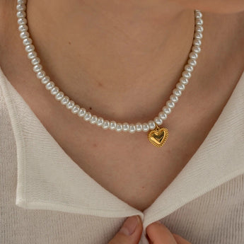 A woman's hand gently touching her neck, wearing an elegant bridal pearl choker with a gold heart charm.