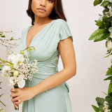 Elegant cream backless evening gown featuring a large bow at the back, worn by a woman standing gracefully, highlighting the dress's flowing silhouette and fitted waist, suitable for wedding guests.
