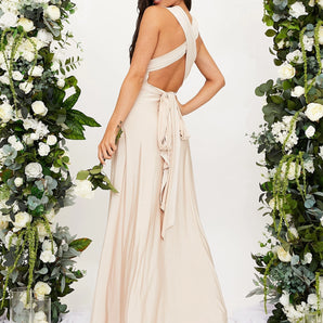 Elegant cream backless evening gown with a bow, featuring floral elements around the shoulder area. The dress has a sophisticated neckline and fitted waist, perfect for a wedding guest. The model is standing, showcasing the dress's graceful design.