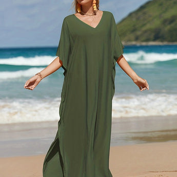 Elegant green V-neck caftan worn by a person standing on a beach with a clear sky and ocean in the background. The lightweight, flowy dress features short sleeves and a cinched waist.