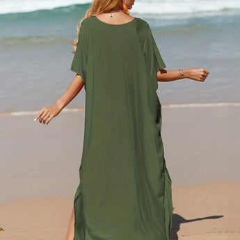 A woman wearing an elegant green V-neck caftan stands on a beach, smiling and enjoying the sunshine. The lightweight, flowy beach dress moves gently with the breeze, showcasing its relaxed fit and comfortable sleeves. The ocean and sand are visible in the background, enhancing the serene, nature-filled setting.
