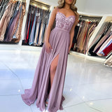 
A woman wearing an elegant lavender lace gown with a sweetheart neckline and high slit, styled in the Viola color and size XL. The dress features intricate lace detailing and is designed with a fitted waist and cap sleeves, perfect for a bridal party or formal event.