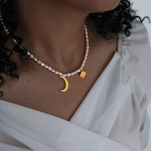 A woman with black hair wears an elegant minimalist pearl choker adorned with a gold moon pendant around her neck.