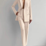 Elegant pink women's suit consisting of a tailored blazer and trouser set, featuring a beige color. The blazer has a structured collar and long sleeves, and the model is standing.