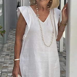 Elegant white linen V-neck summer dress worn by a smiling woman accessorized with a pearl necklace, sunglasses, and a fashionable handbag.