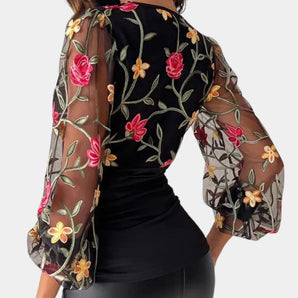 Elegant black blouse featuring sheer sleeves with intricate floral embroidery, highlighting the neckline and waist area, worn by a model displaying the blouse's design.