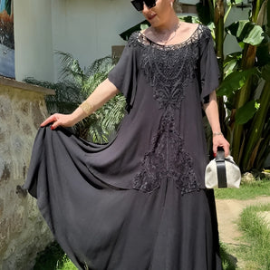 Embroidered black lace maxi dress in a bohemian style, featuring long sleeves and a fitted waist. The gown is displayed outdoors, with grass and plants in the background, creating a summery, elegant look suitable for formal occasions.