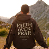 A person wearing a Faith Over Fear Hoodie in coffee color, size 5XL, standing outdoors with a natural environment backdrop including plants and mountains.
